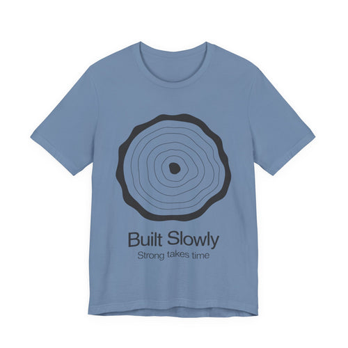 Blue t-shirt with black circular design and text 'Built Slowly Strong takes time' on a white background