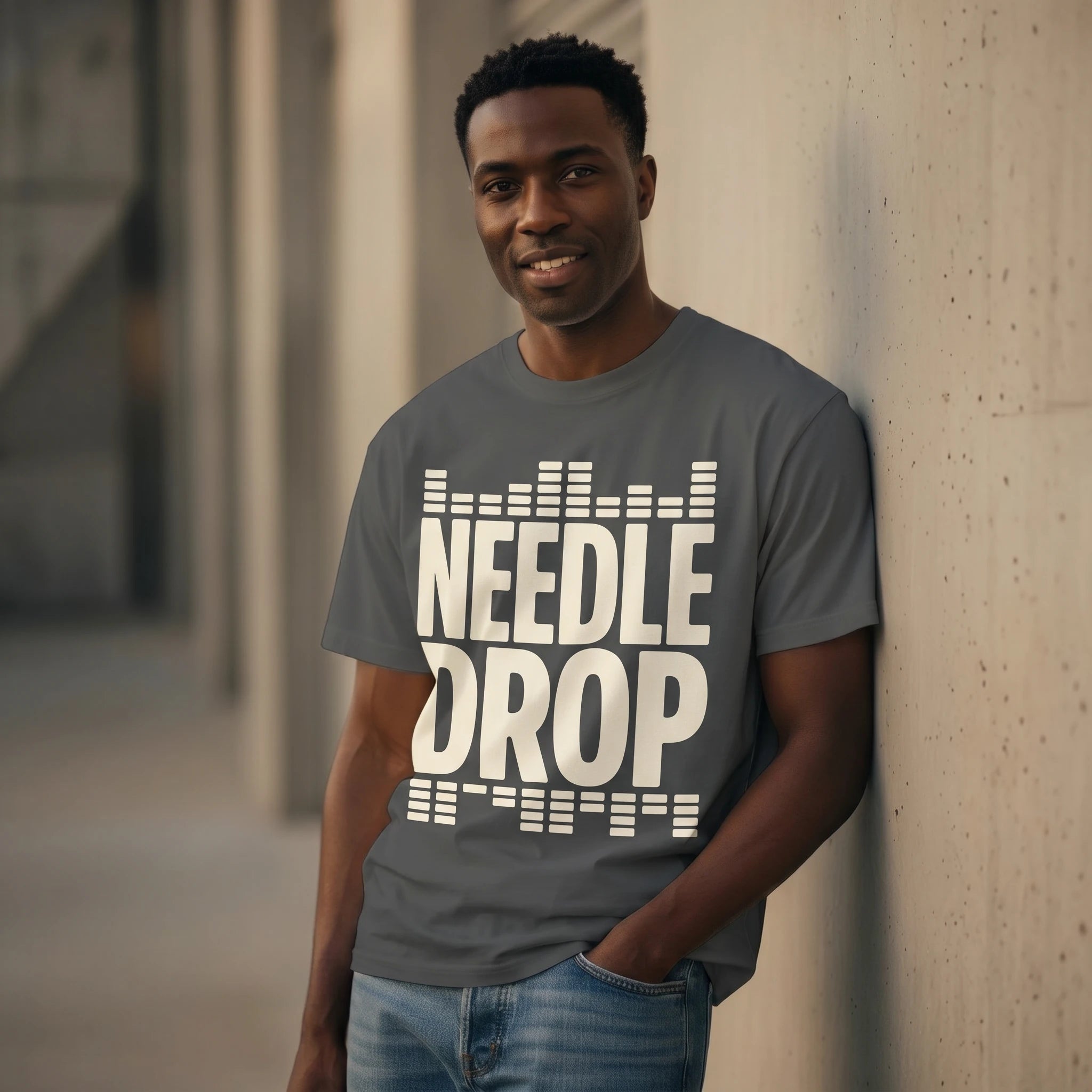 Man wearing a gray t-shirt with 'Needle Drop' text, leaning against a wall.