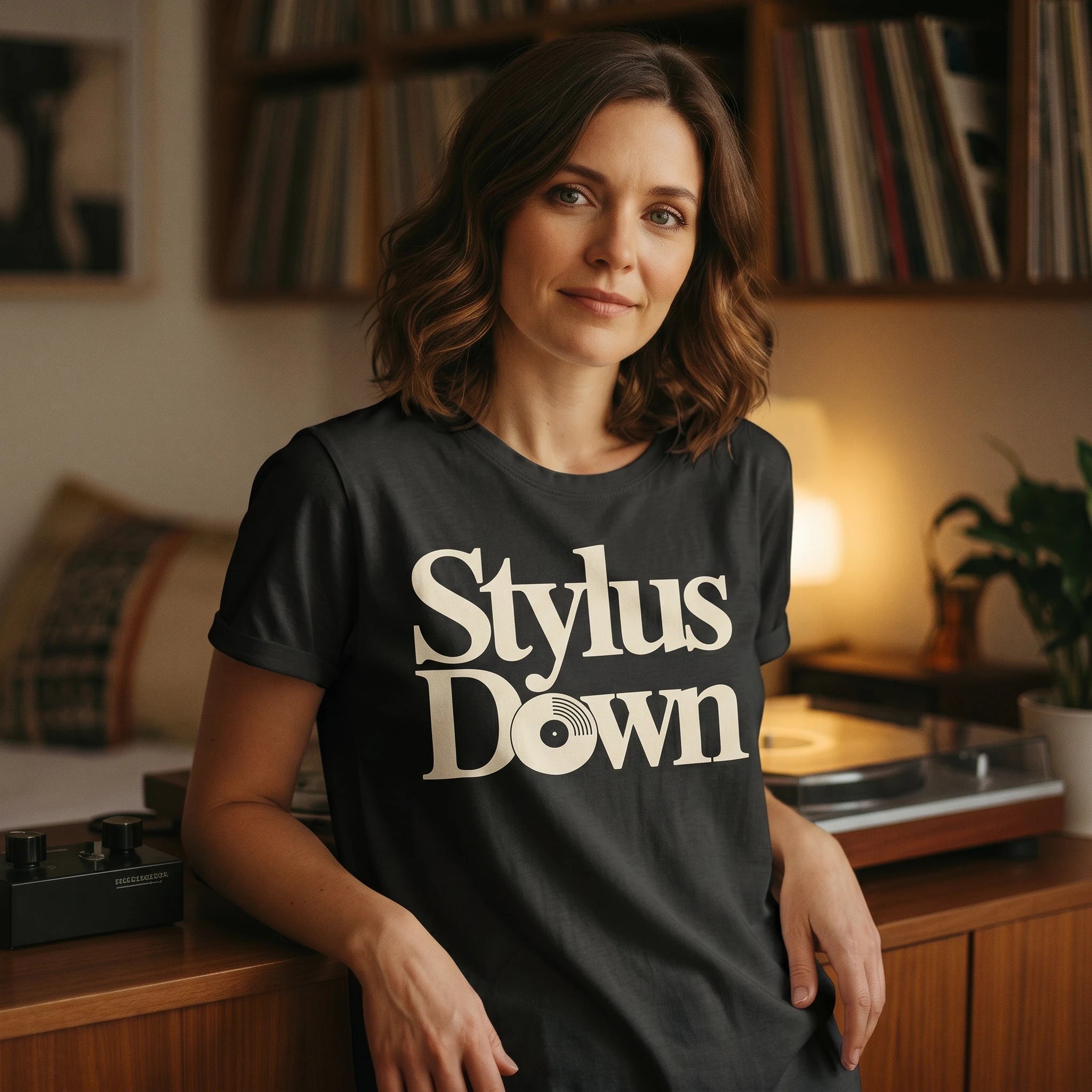 Woman wearing a black t-shirt with 'Stylus Down' text in a room with records and a lamp.