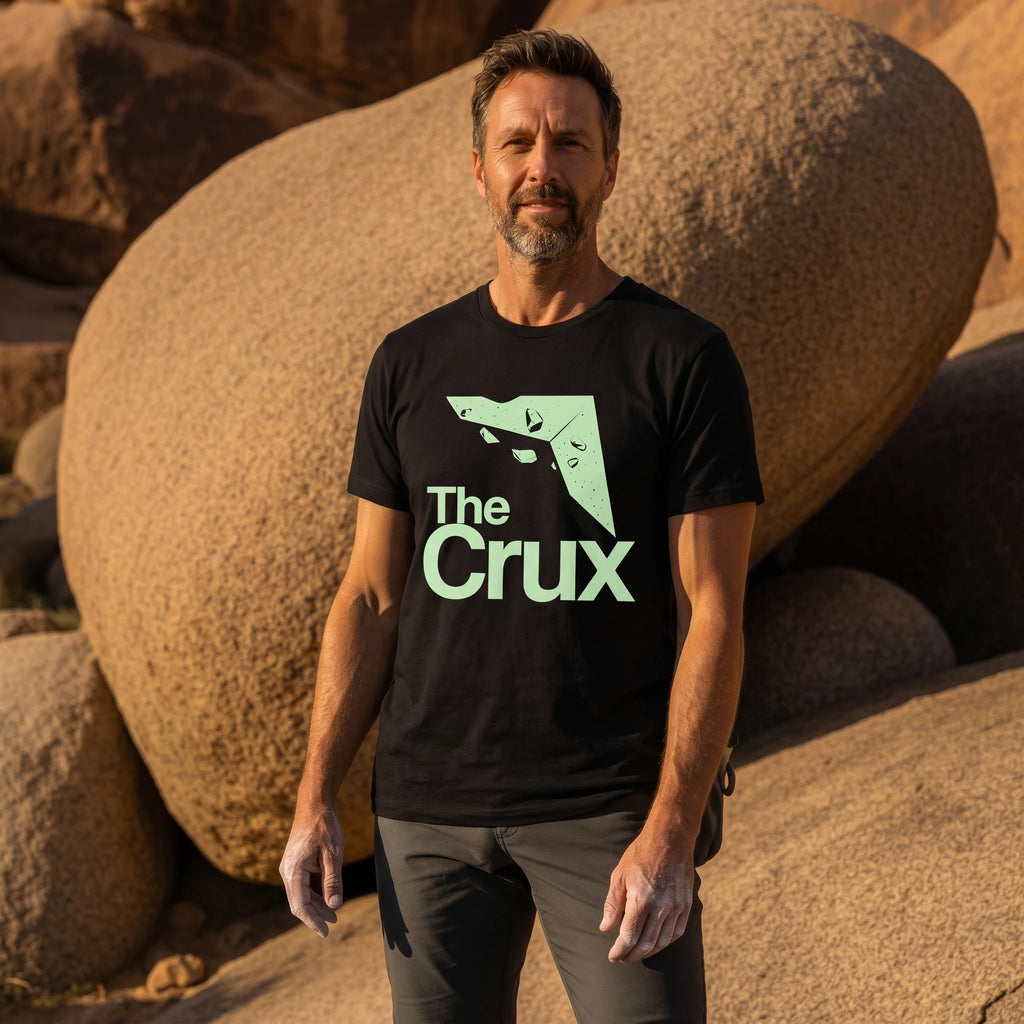 Man wearing a black t-shirt with 'The Crux' logo standing in front of large rocks.