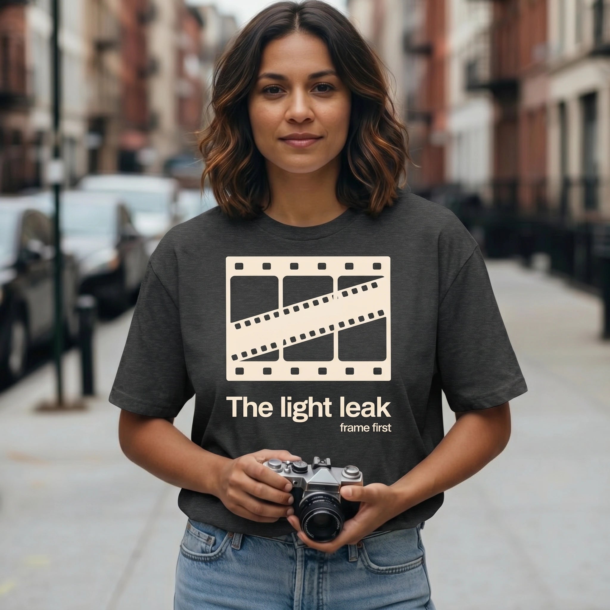 Person wearing a t-shirt with 'The light leak frame first' design, holding a camera on a city street.
