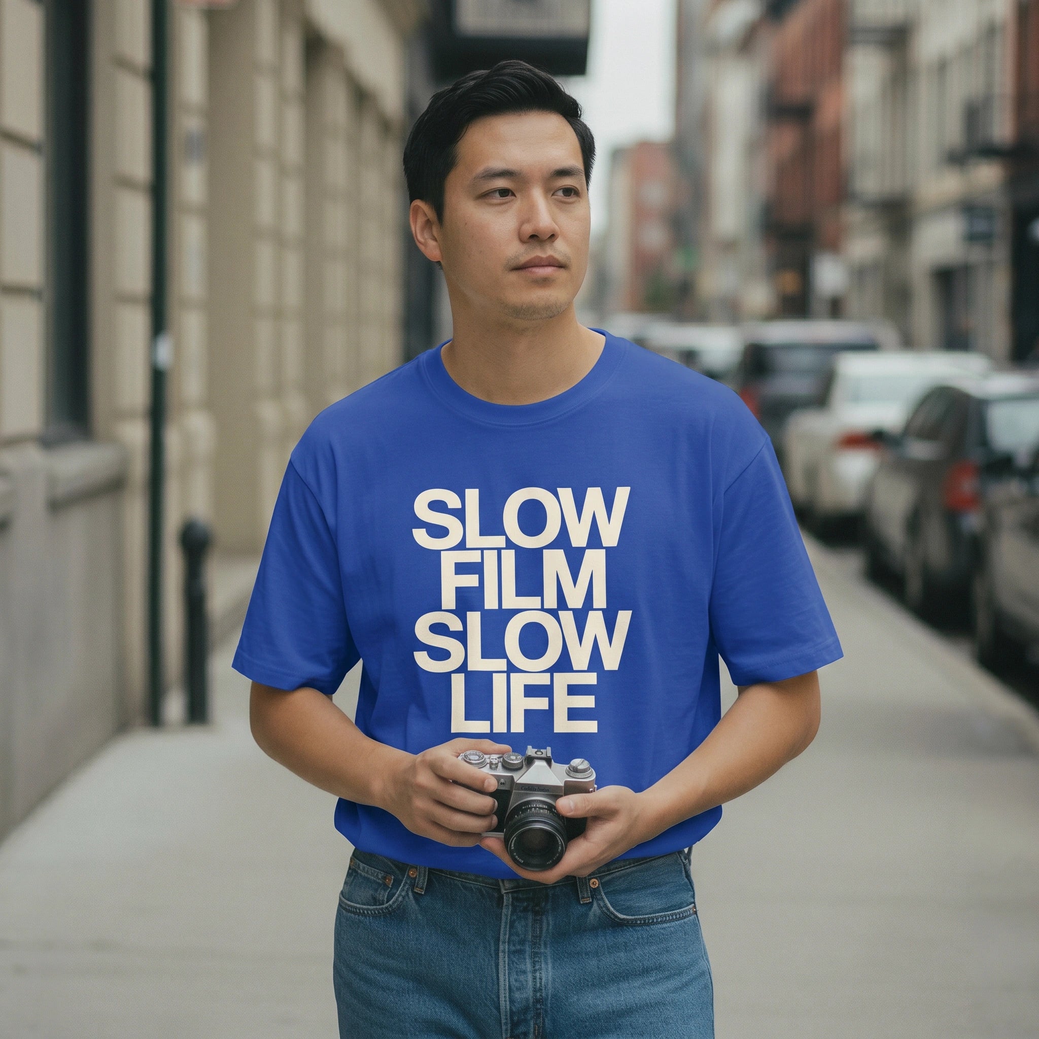 Man wearing a blue t-shirt with 'SLOW FILM SLOW LIFE' text, holding a camera on a city street.