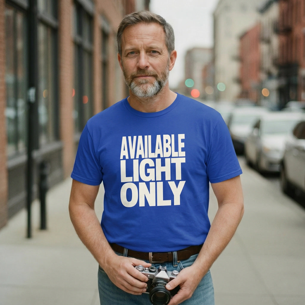 Man wearing a blue t-shirt with 'AVAILABLE LIGHT ONLY' text, holding a camera on a city street.