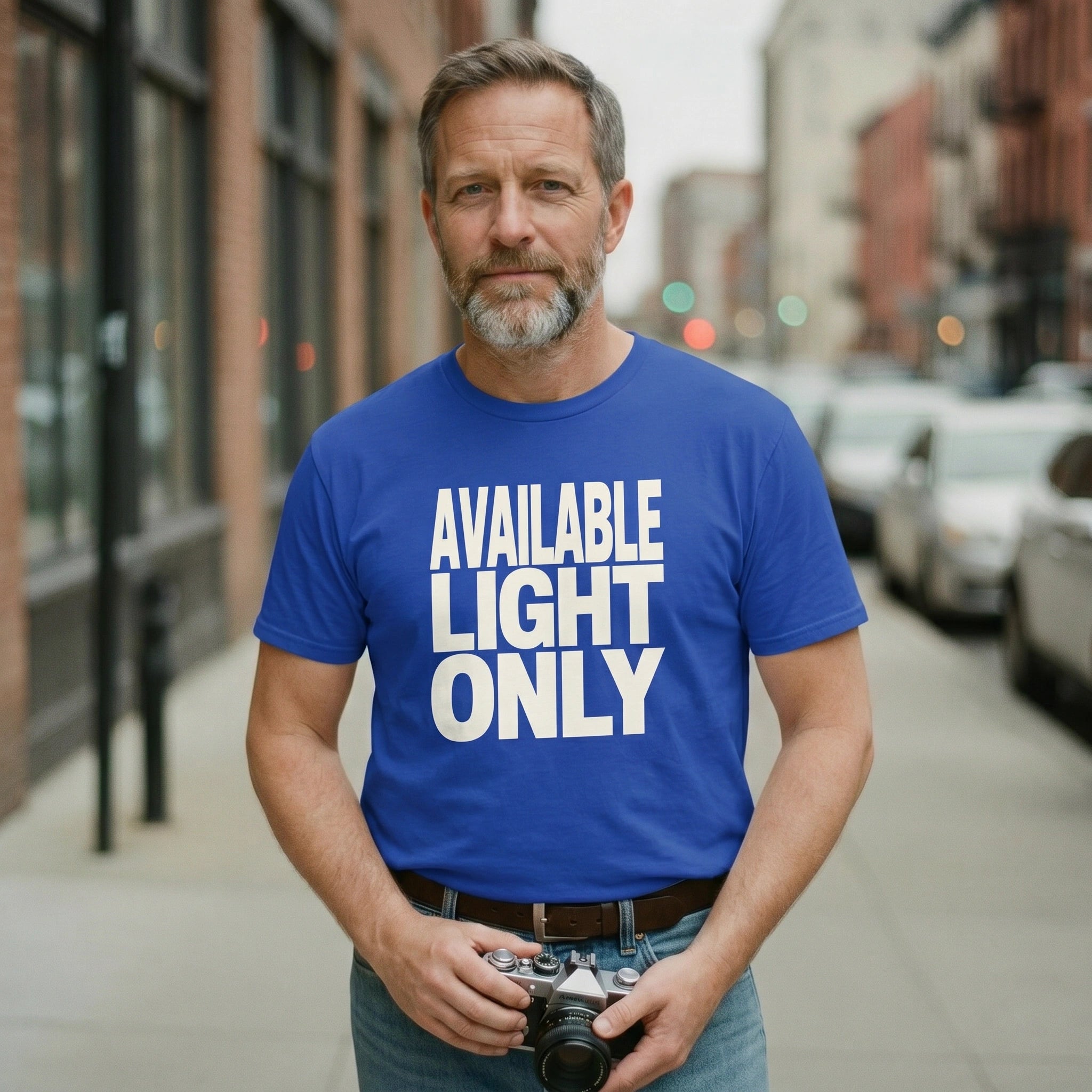 Man wearing a blue t-shirt with 'AVAILABLE LIGHT ONLY' text, holding a camera on a city street.