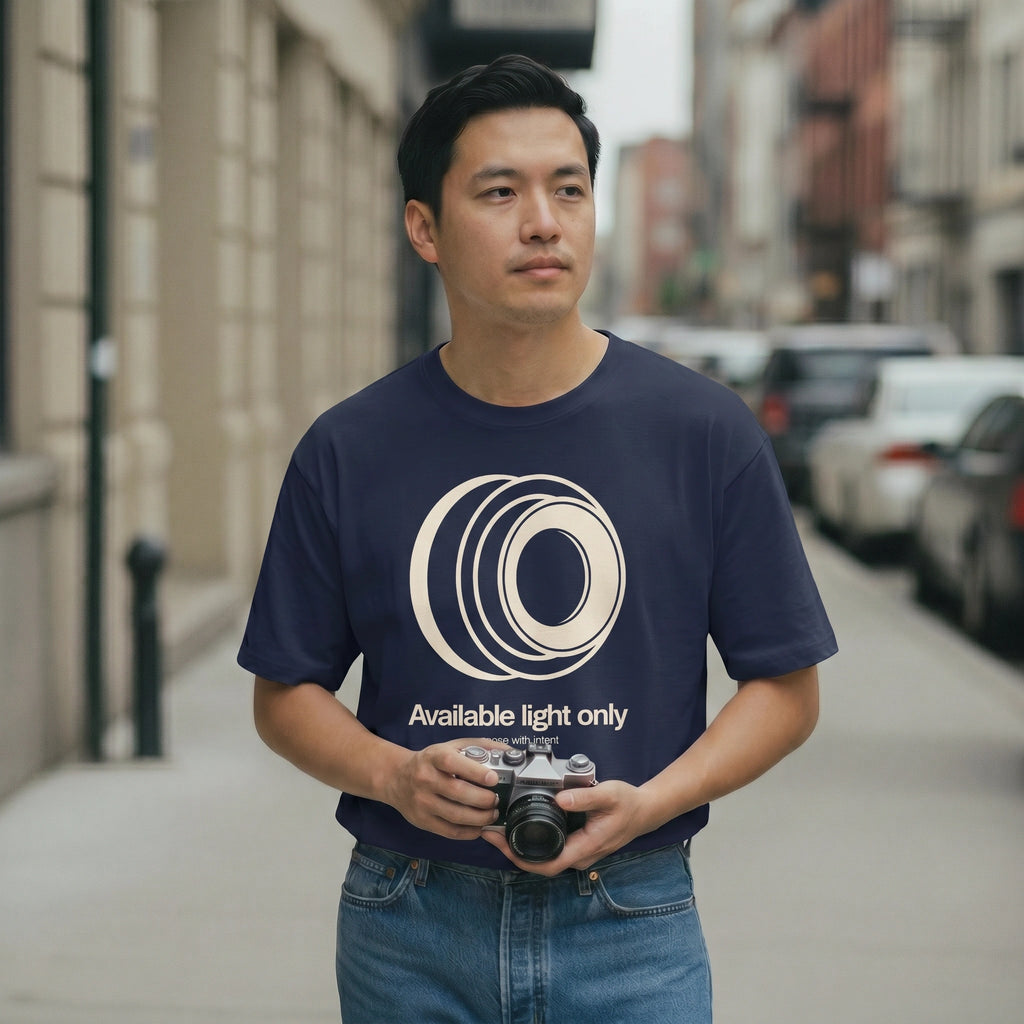 Man wearing a navy blue t-shirt with a camera graphic and text, holding a camera on a city street.