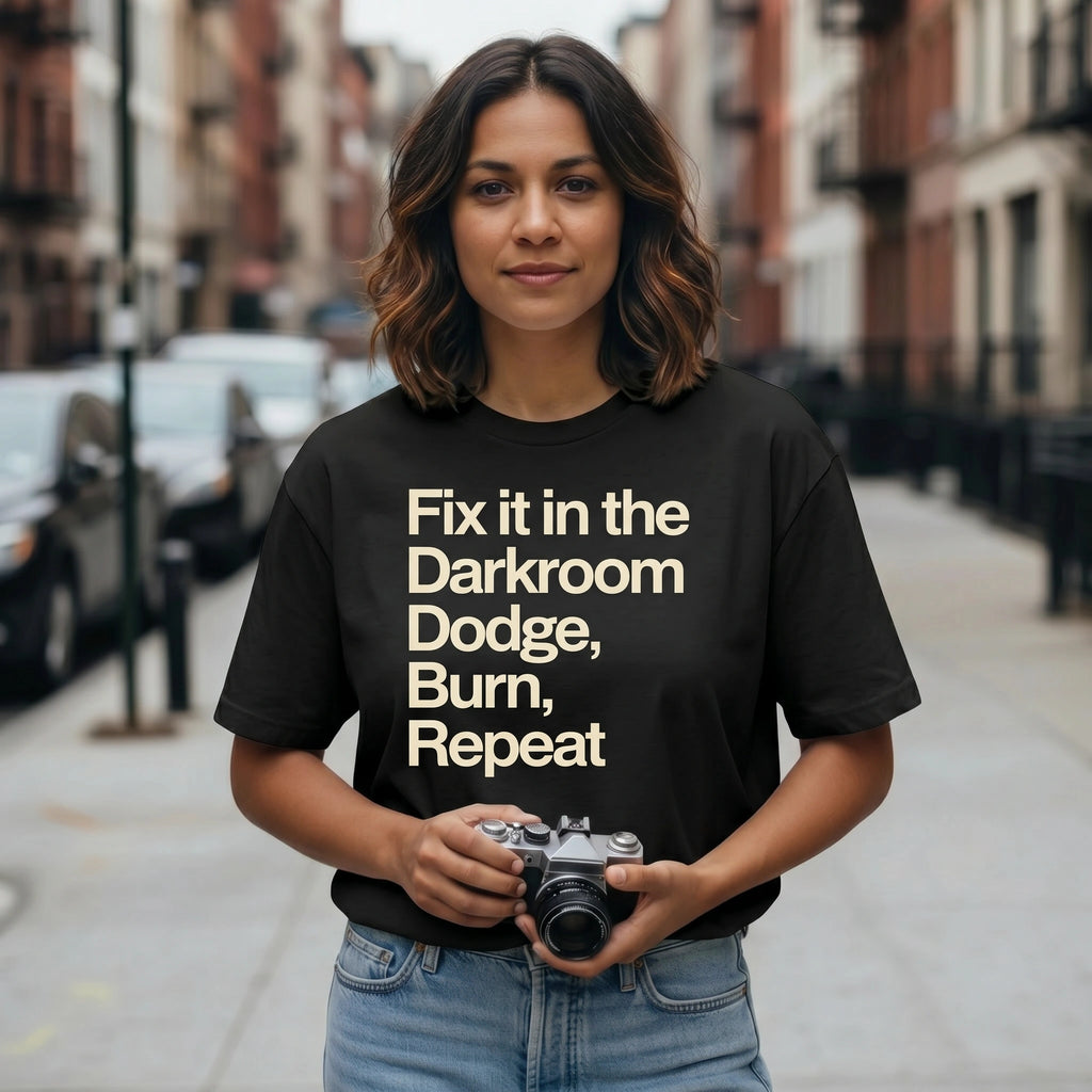 Person wearing a black t-shirt with 'FIX IT IN THE DARKROOM' text holding a camera on a city street.
