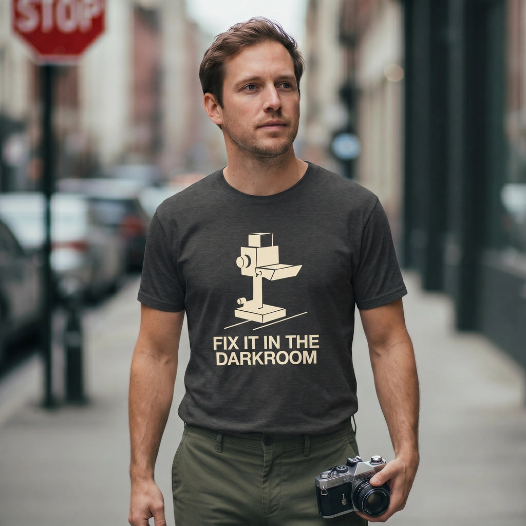 Man wearing a t-shirt with a darkroom graphic and 'FIX IT IN THE DARKROOM' text, holding a camera on a city street.