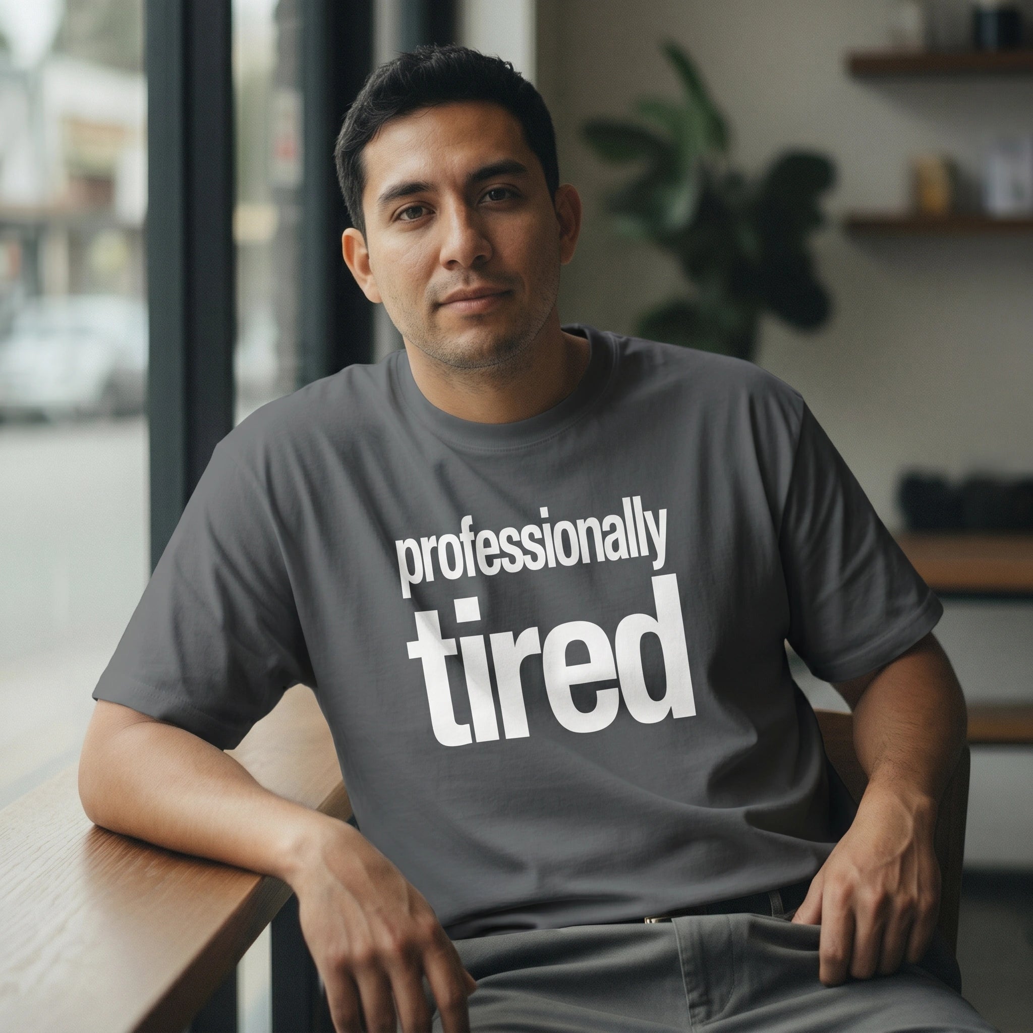 Man wearing a gray t-shirt with 'professionally tired' text, sitting indoors.