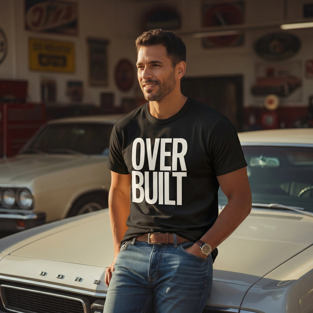 Man wearing a 'OVERBUILT' t-shirt standing next to a classic car in a garage.