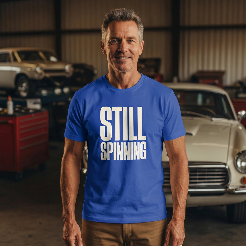 Man wearing a blue 'STILL SPINNING' t-shirt in a garage with vintage cars.
