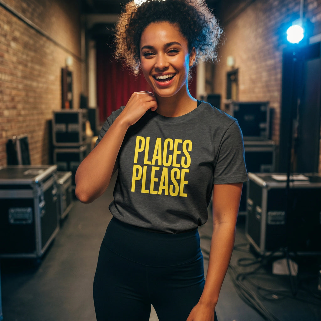 Person wearing a gray t-shirt with 'PLACES PLEASE' in yellow text, standing in a room with equipment.