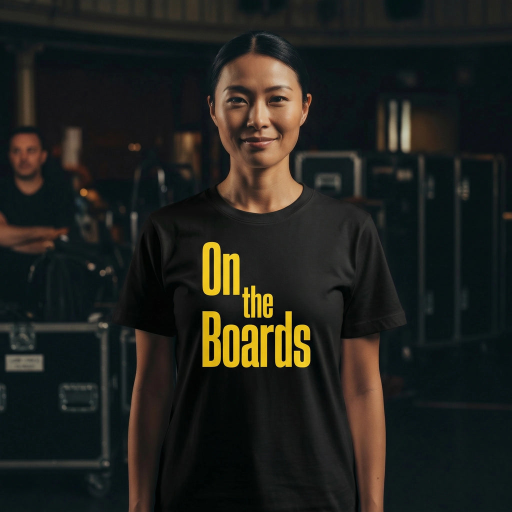 Person wearing a black t-shirt with 'On the Boards' in yellow text, standing in a backstage area.